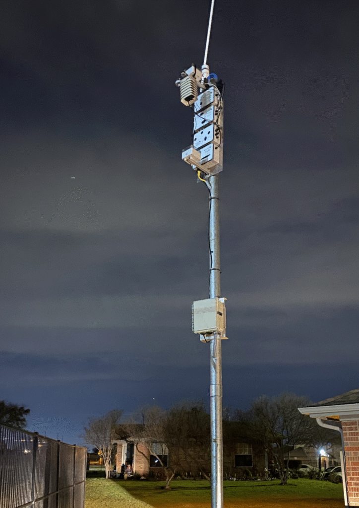 A tall metal pole at night equipped with multiple air-monitoring devices and sensors, standing in a residential yard with houses and trees visible under a cloudy sky.