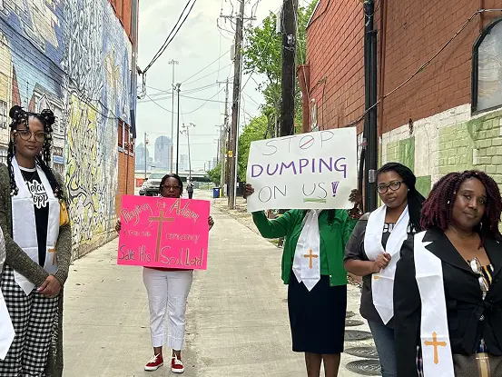 A group of community members standing in an alleyway during a protest, some wearing white stoles with crosses. Several people hold handmade signs, including one that reads “STOP DUMPING ON US!” and another about saving a local community. Colorful murals and brick buildings line the alley behind them.